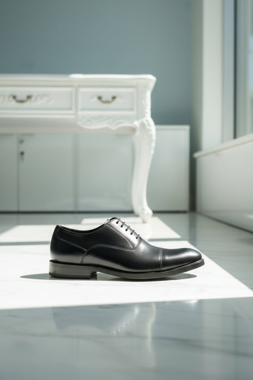 A black leather lace-up dress shoe displayed on a teal velvet bench inside a shoe store, with shelves of shoes and seating visible in the background.
